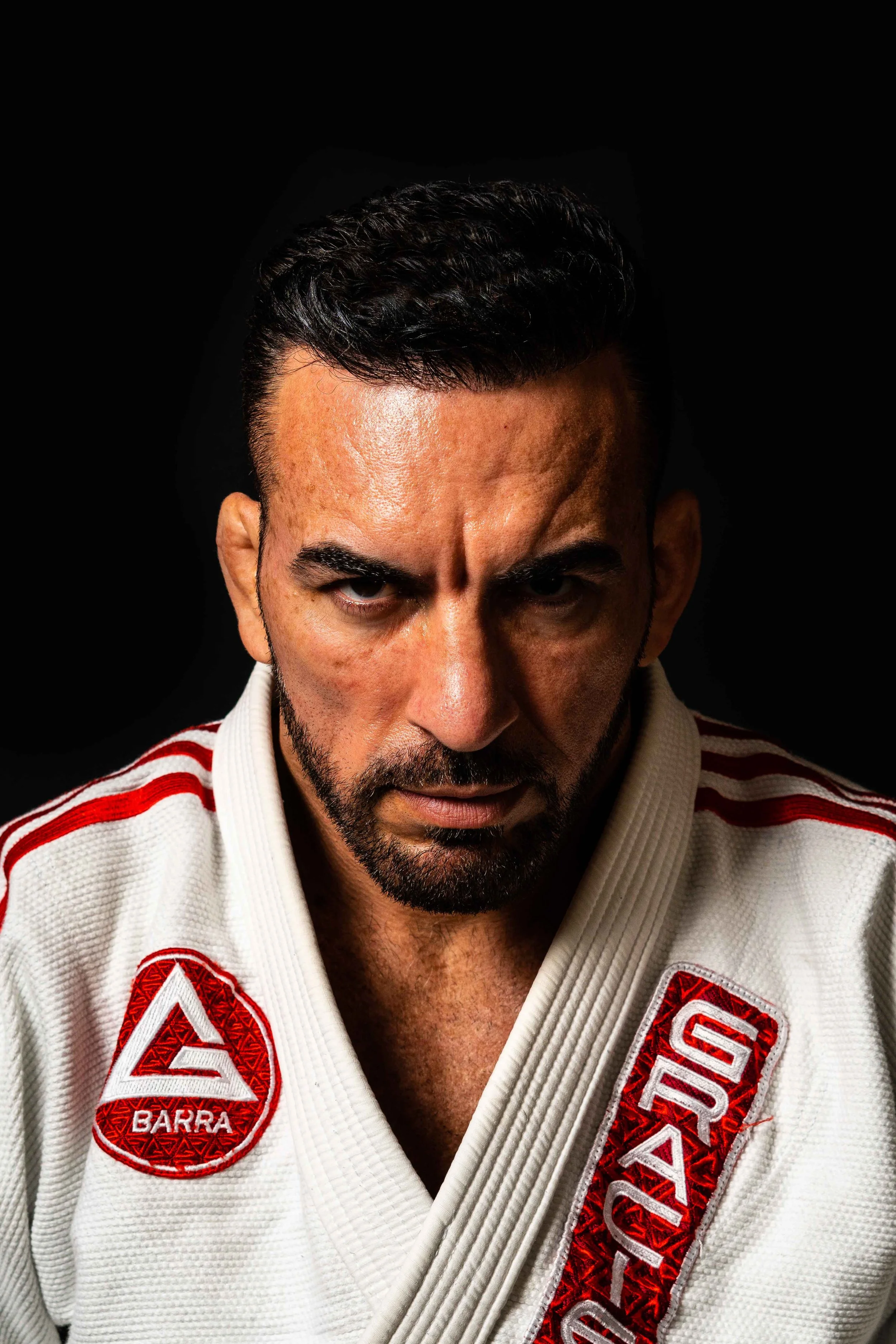 A man with dark hair and a beard, wearing a white martial arts gi with red and white patches, looking directly at the camera against a black background.
