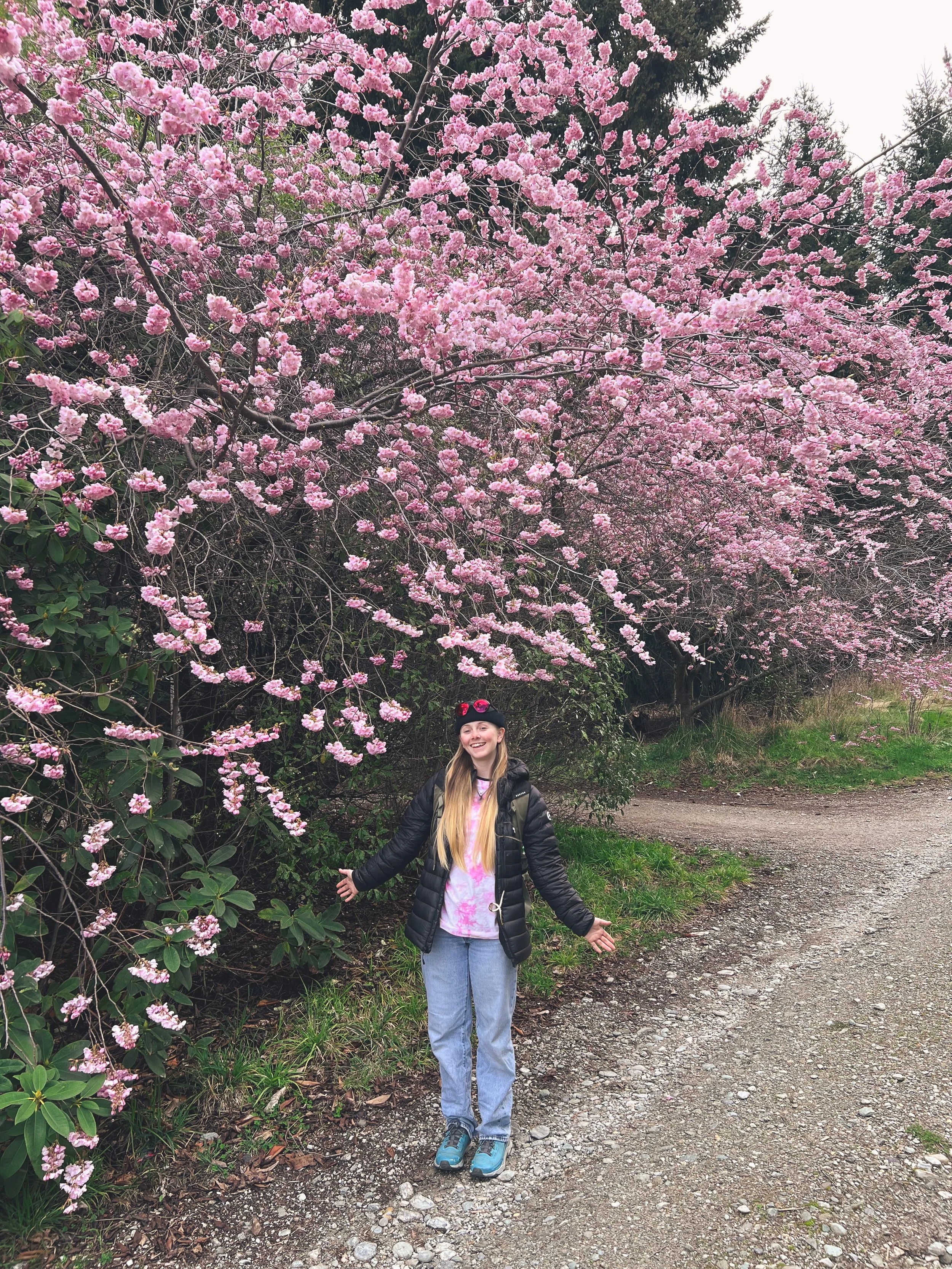 A young woman standing on a dirt path near pink blossoming trees, smiling with her arms slightly outstretched.