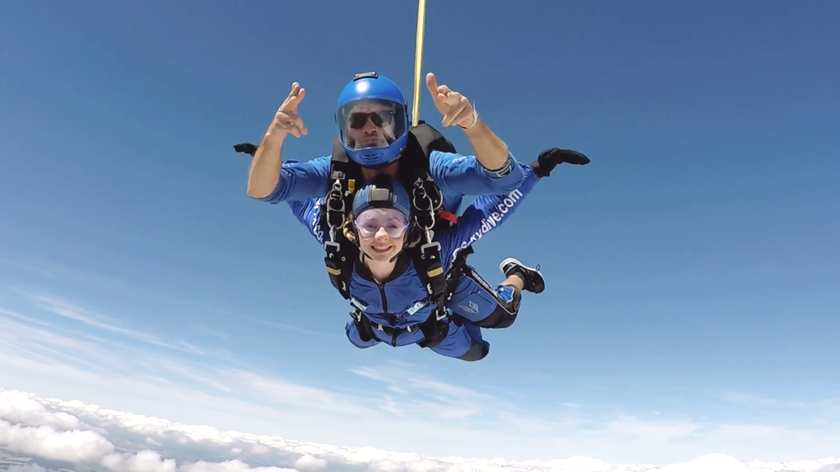 Two people tandem skydiving in the sky above clouds, wearing blue jumpsuits and helmets, with one person smiling at the camera.
