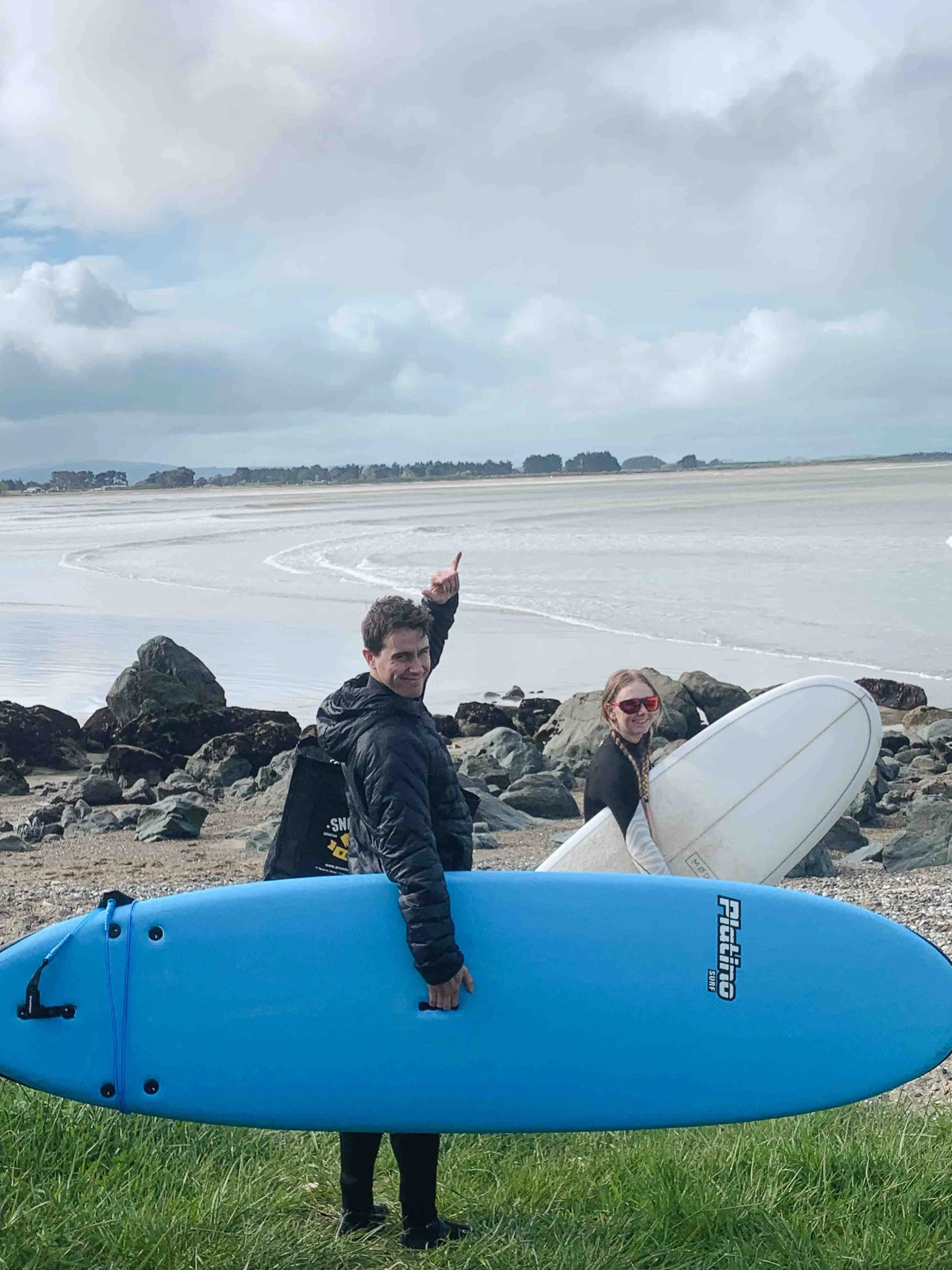 Two people standing outdoors near rocks with large blue and white surfboards. The background features a cloudy sky over the ocean and beach