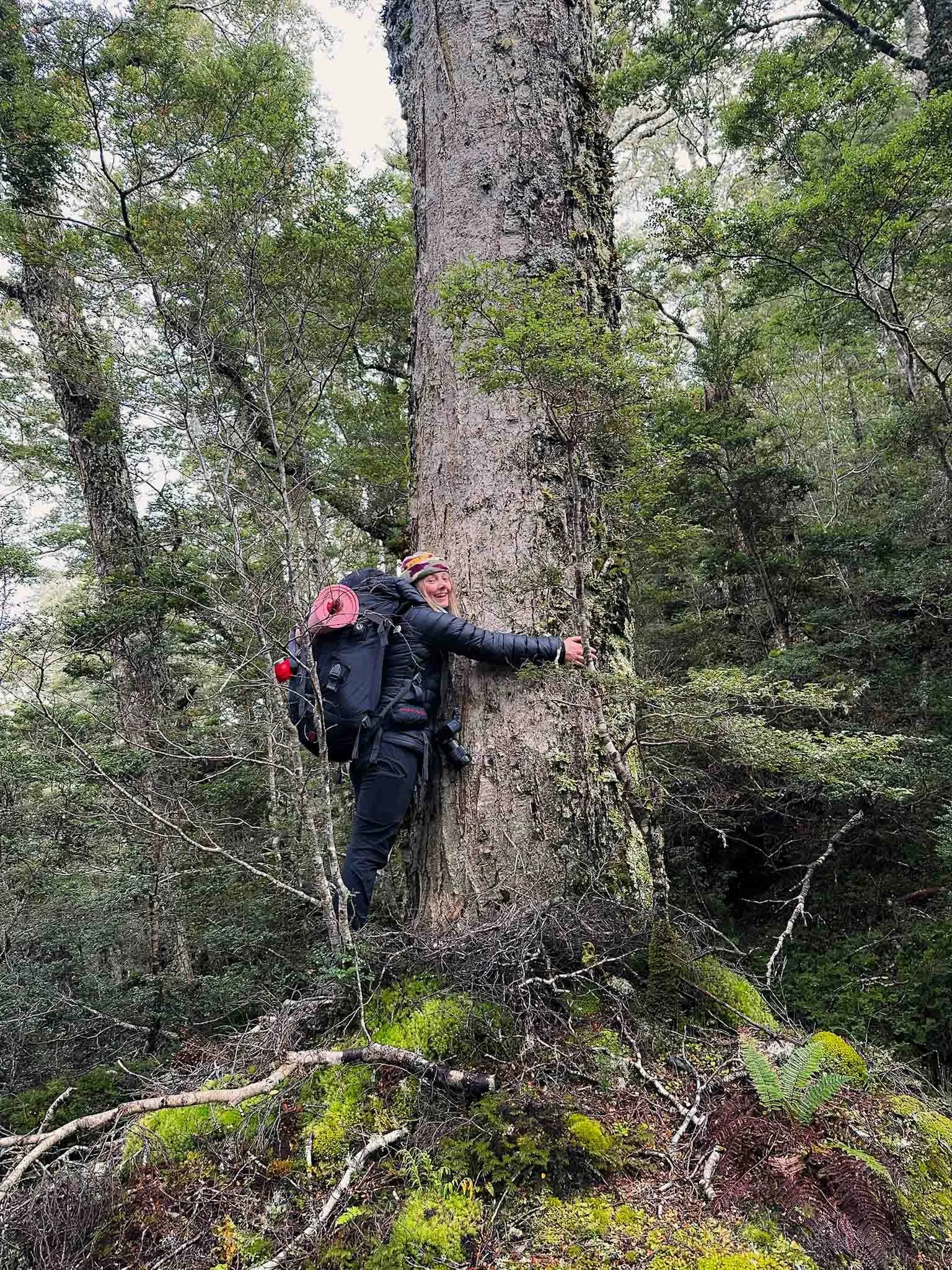 A woman with a backpack and helmet standing next to a large tree trunk in a dense forest, hugging the tree and smiling.