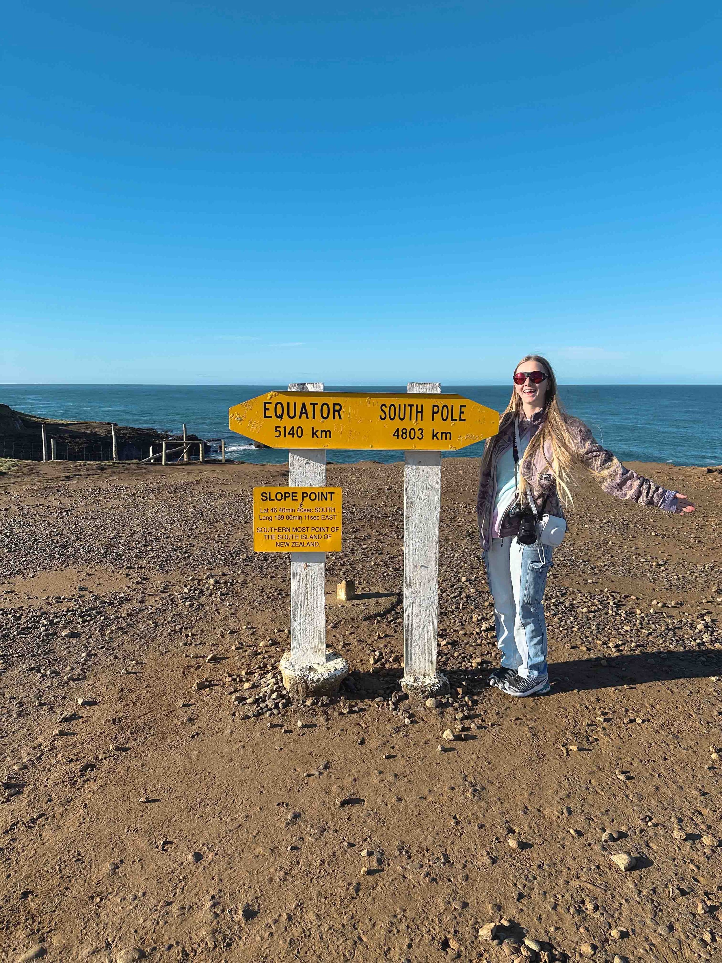 A woman with long blonde hair wearing sunglasses, a patterned jacket, and jeans standing next to a bright yellow sign at the South Point of New Zealand's South Island, with the ocean and a clear blue sky in the background.