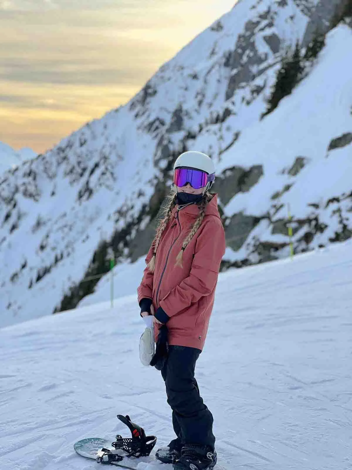 A person in pink winter gear and a white helmet standing on snow with their snowboard attached on one foot, mountains in the background, and a colorful sunset sky.