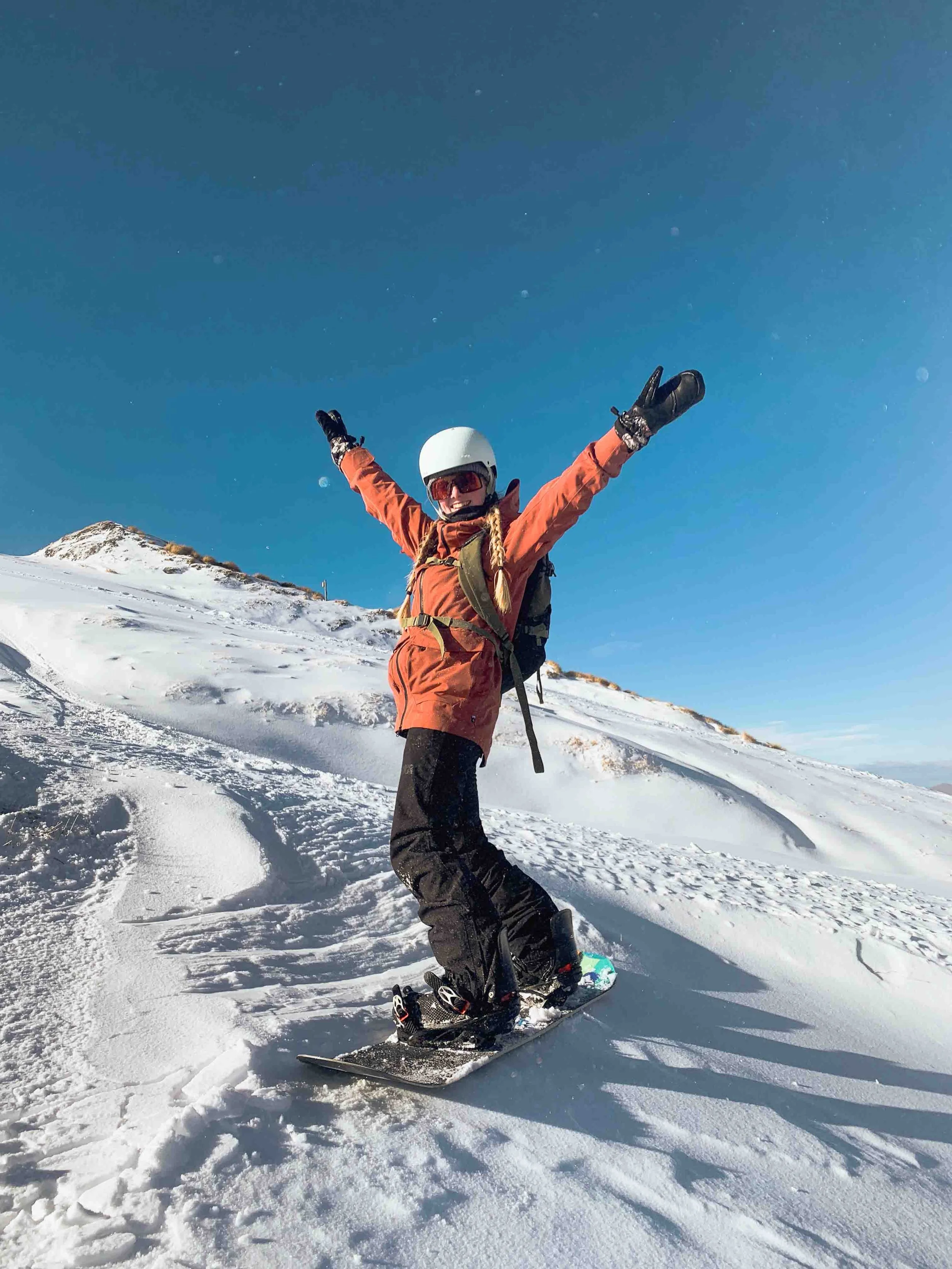 A person standing on snow with arms raised, wearing snowboarding gear including a helmet, goggles, and winter clothing, on a snowy mountain under a clear blue sky.