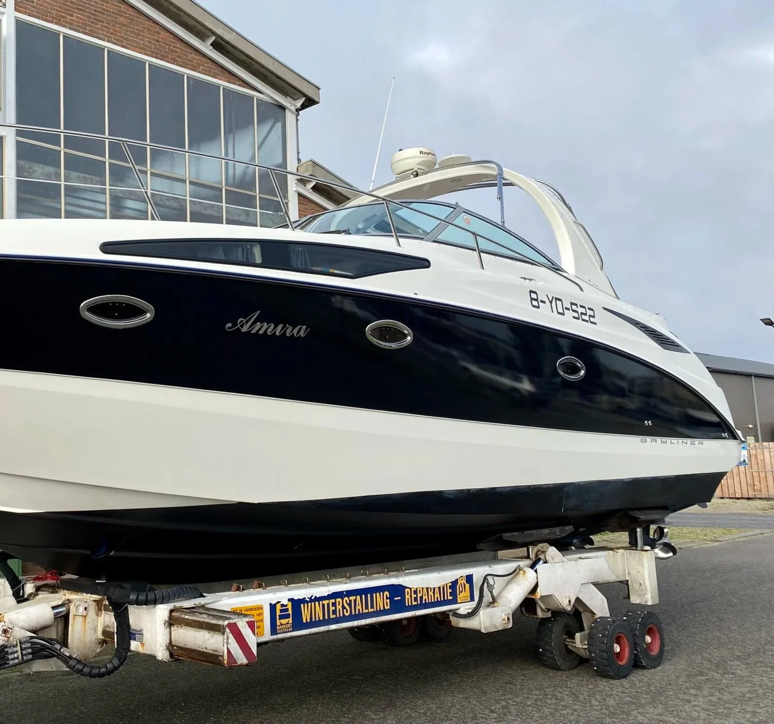 A white and black yacht named Amura on a trailer with the license plate 8-YO-522 parked on a street near a modern building. The trailer has a sign that reads 'Winterstalling - Repareratie' and is equipped with wheels and black hoses.