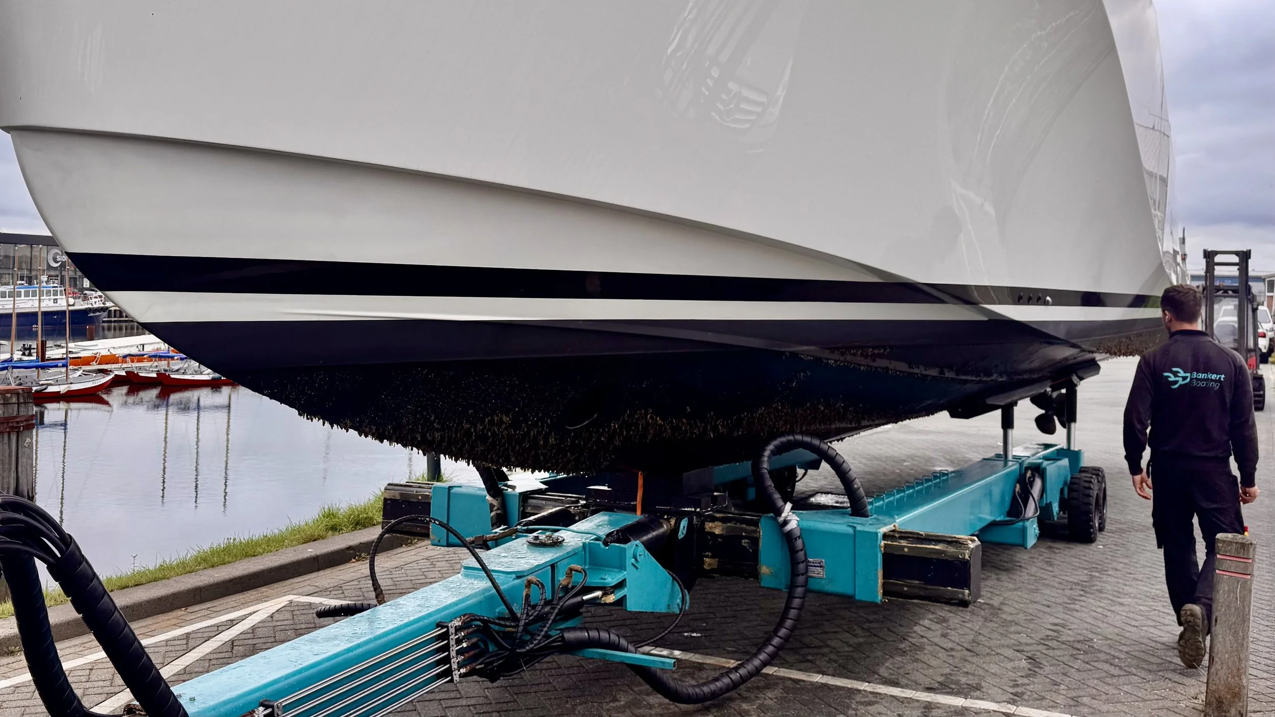 Large white yacht on a teal support with a person walking beside it near a harbor with smaller boats and water.