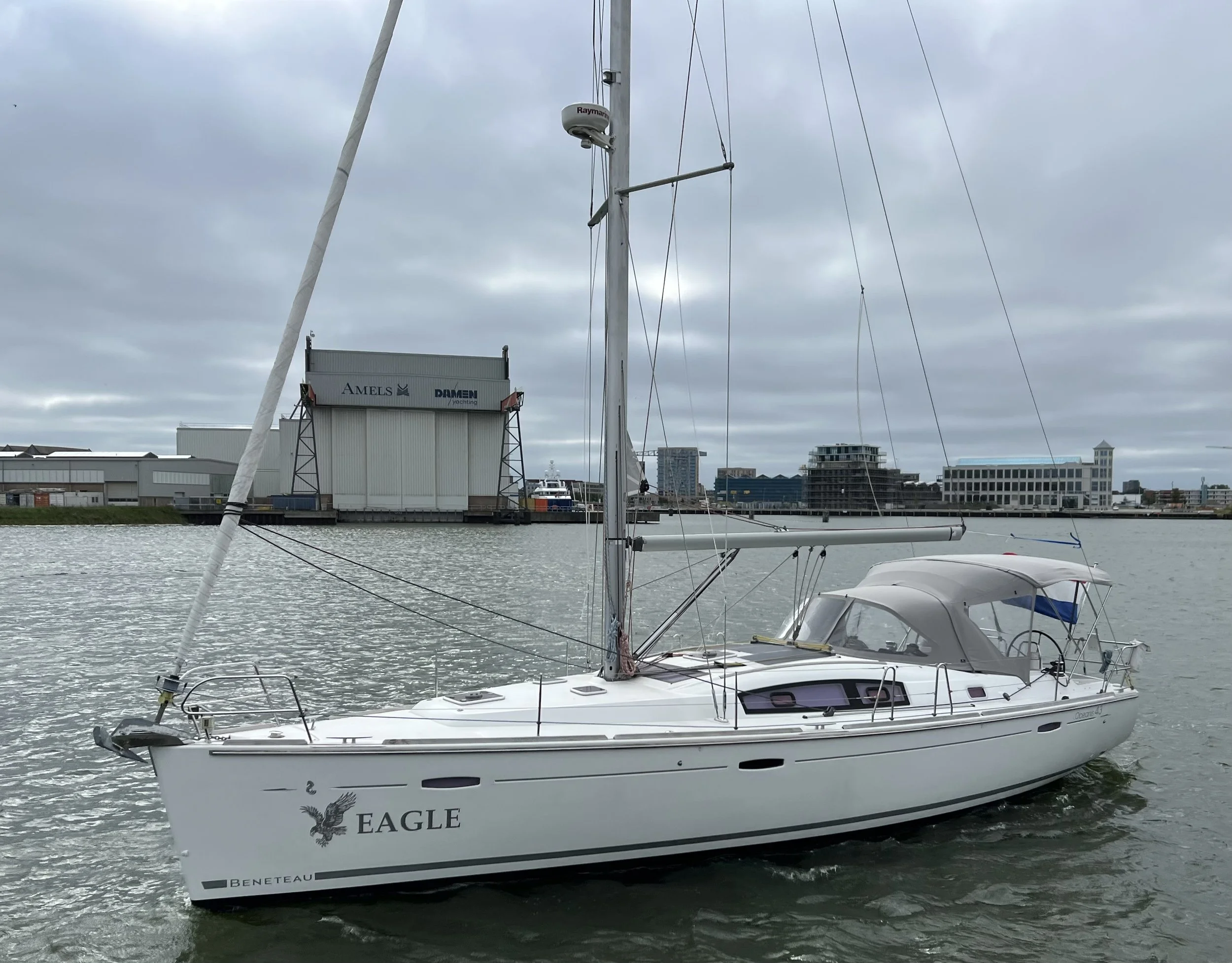 White sailboat named Eagle with a covered cockpit, moored in a harbor, with industrial buildings and city skyline in the background under cloudy sky.