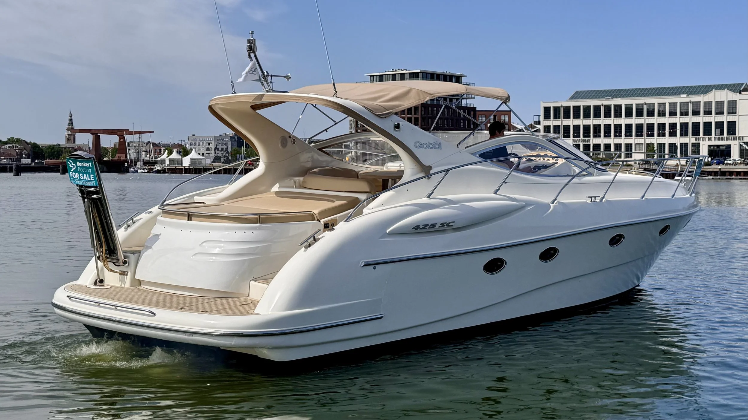 A white motor yacht with beige seating and a beige canopy, cruising on a body of water with a city skyline and buildings in the background.