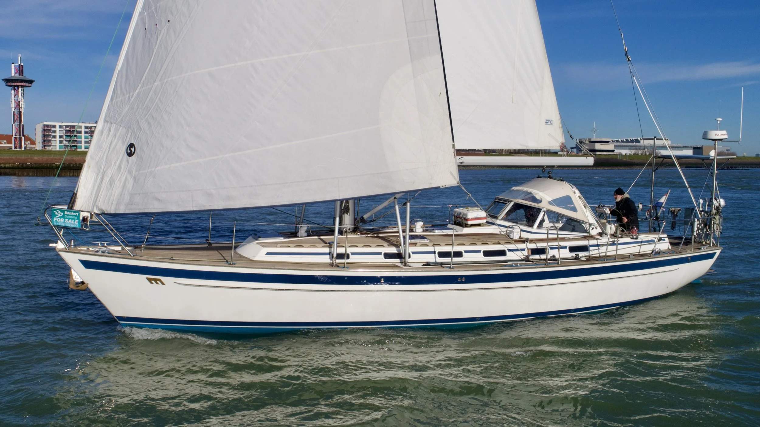 A white sailboat with a large wind-filled sail on a body of water, with buildings and a lighthouse in the background.