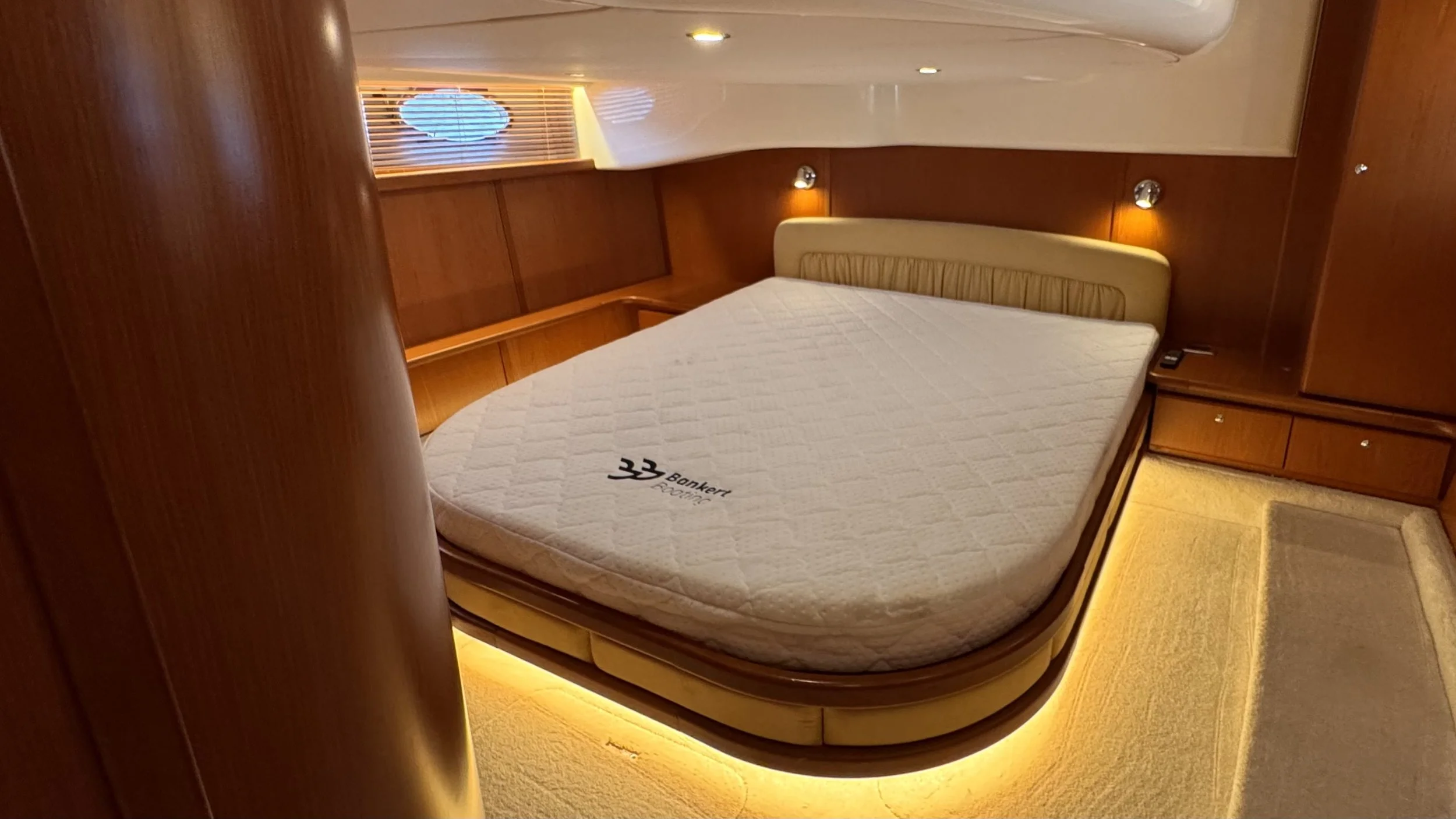 A luxurious bedroom with a round bed, wooden walls, and built-in lighting. The bed has a mattress with the brand name 'Bankert' on it. There is a small bedside table with a remote control on it, and a curved ceiling with small recessed lights. A wind