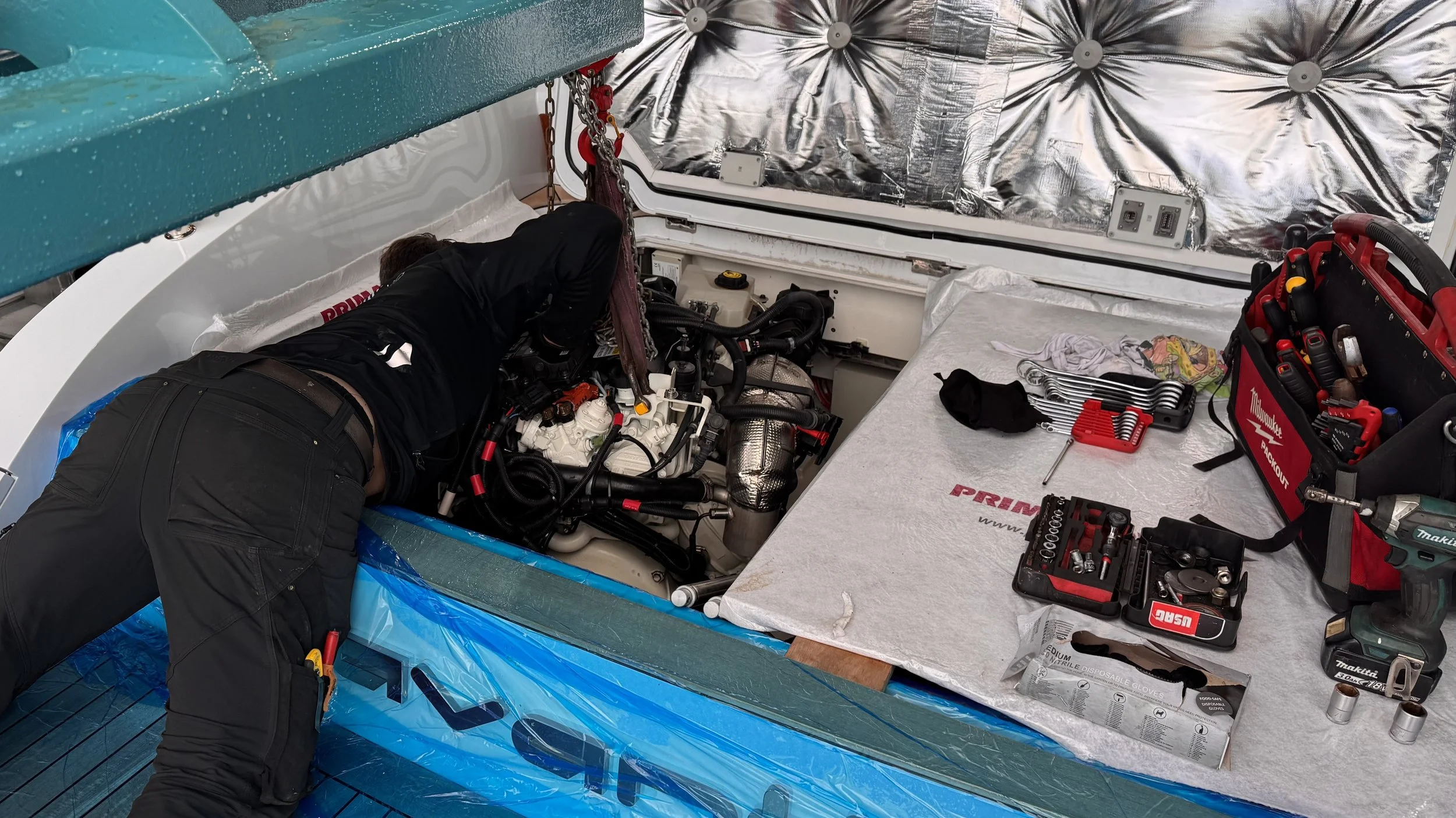 A mechanic working on the engine of a racing boat, which is in a boat shop. The mechanic is bent over the engine, and various tools and toolkits are on a table nearby.