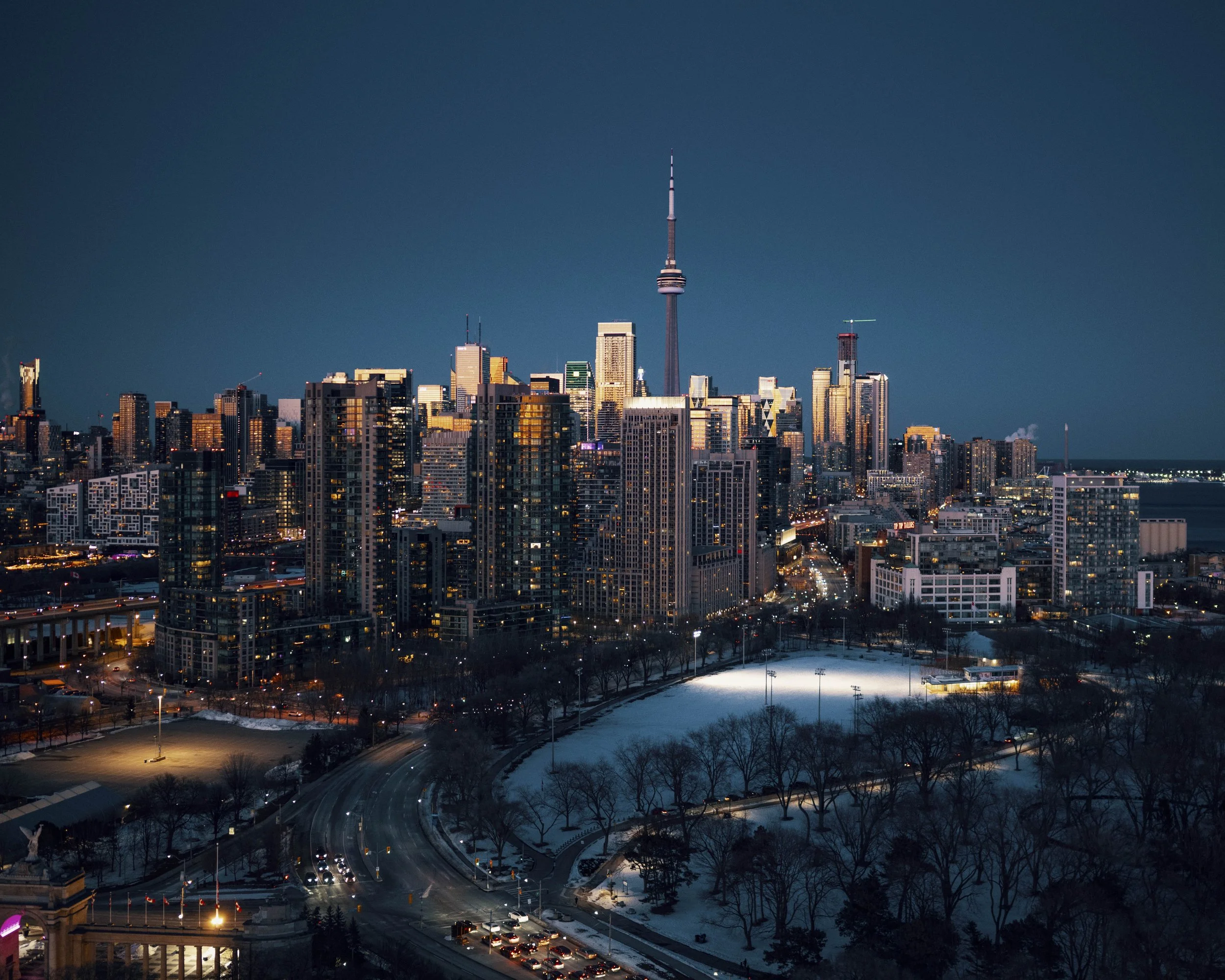 Night view of downtown Toronto with the CN Tower illuminated, surrounded by tall buildings and a snow-covered park in the foreground.