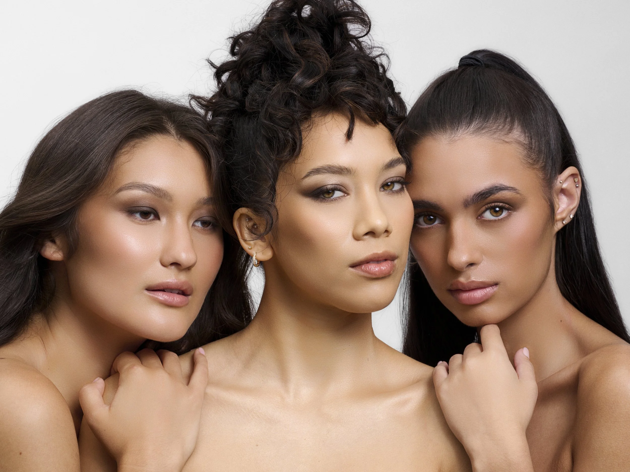 Three women with natural makeup and dark hair posing closely together against a plain white background.