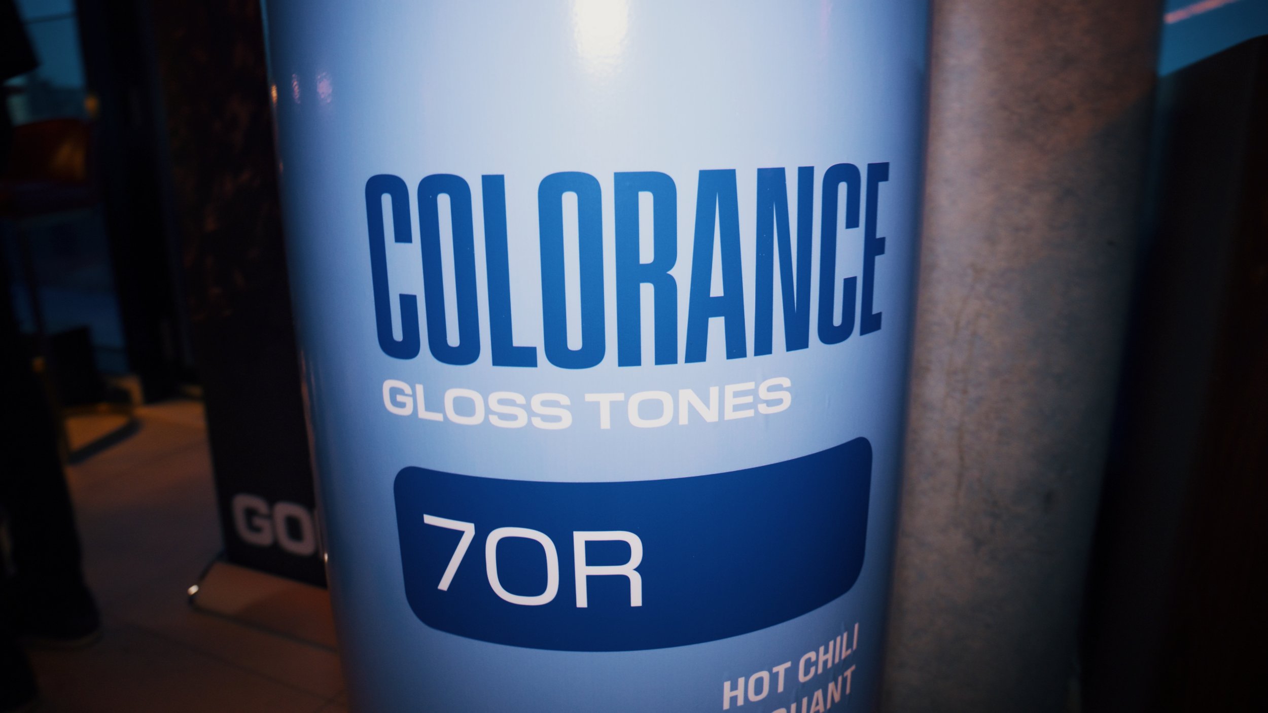 Close-up of a cylindrical container labeled 'COLORANCE GLOSS TONES 70R' with additional text 'HOT CHILI' visible at the bottom, placed outdoors near a wall.