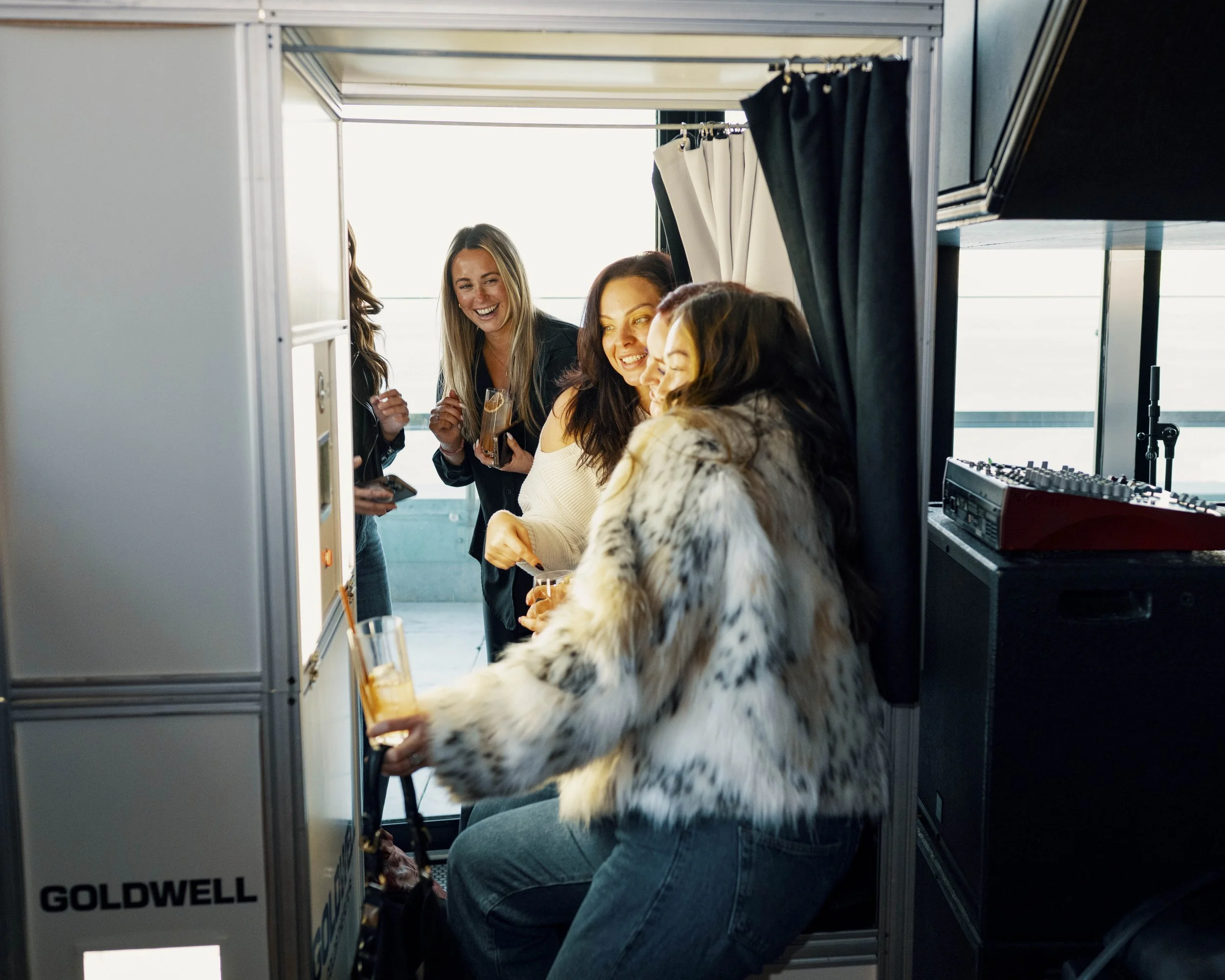 Group of women socializing with drinks in a photobooth on a rooftop terrace, with a DJ setup nearby and city skyline in the background.