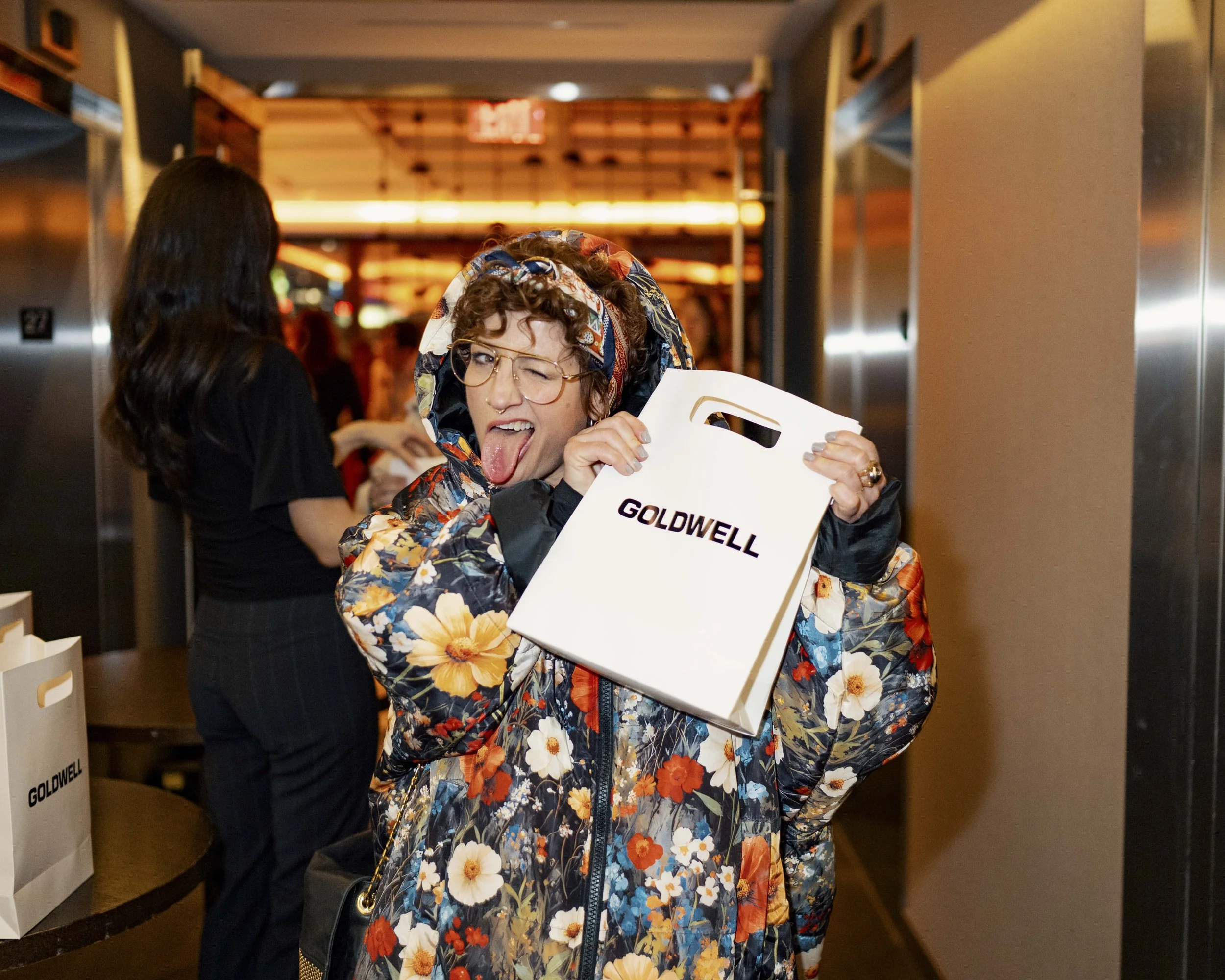 A woman with glasses and curly hair making a playful face with her tongue out while holding a Goldwell shopping bag. She is wearing a floral-patterned jacket and standing inside a mall or shopping center.