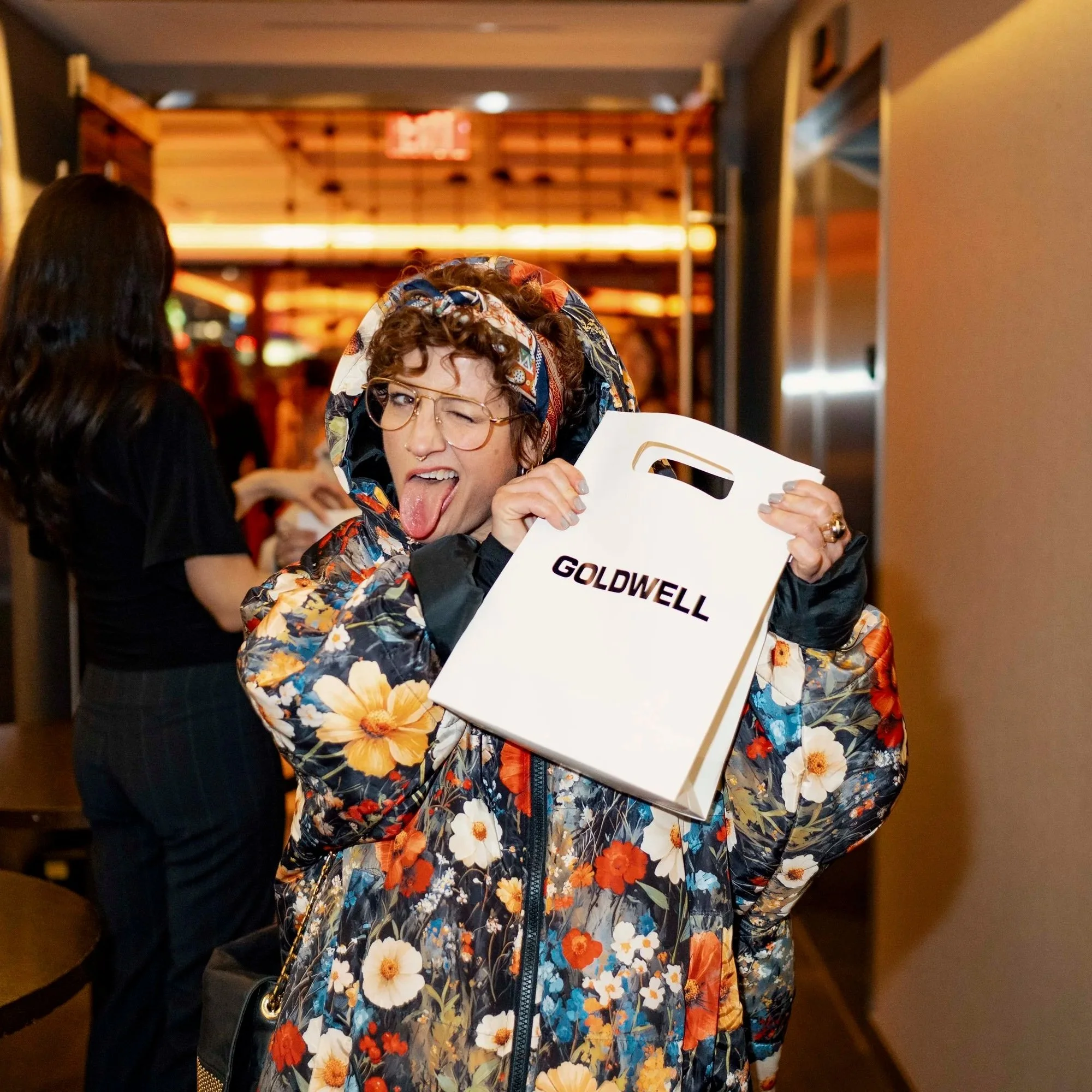 Woman in a floral jacket holding a Goldwell shopping bag, sticking her tongue out and winking, in a lively indoor setting.