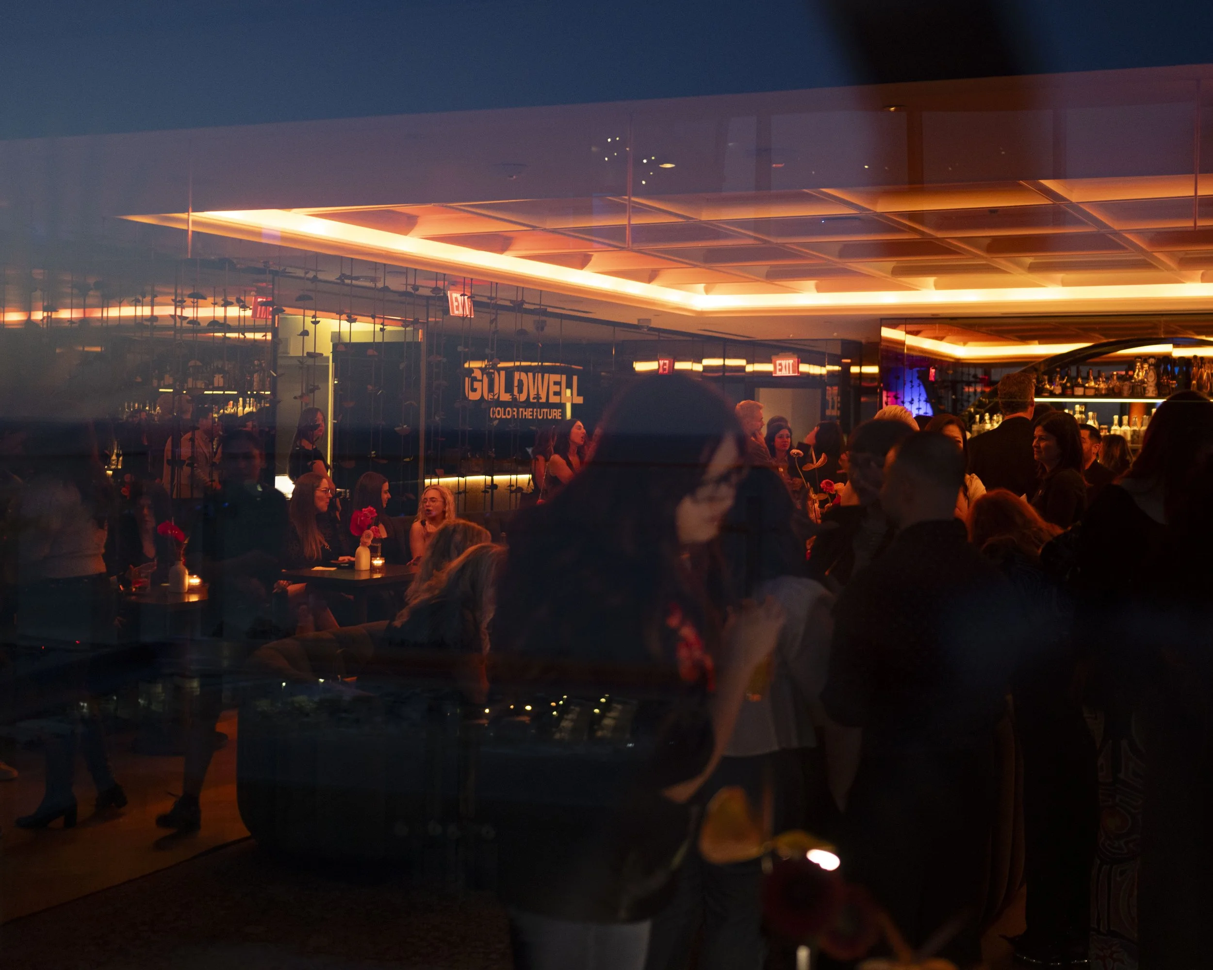 Crowded indoor bar or restaurant at night with people socializing, illuminated by warm lighting, with a reflected sign that reads 'GOLDWELL'.