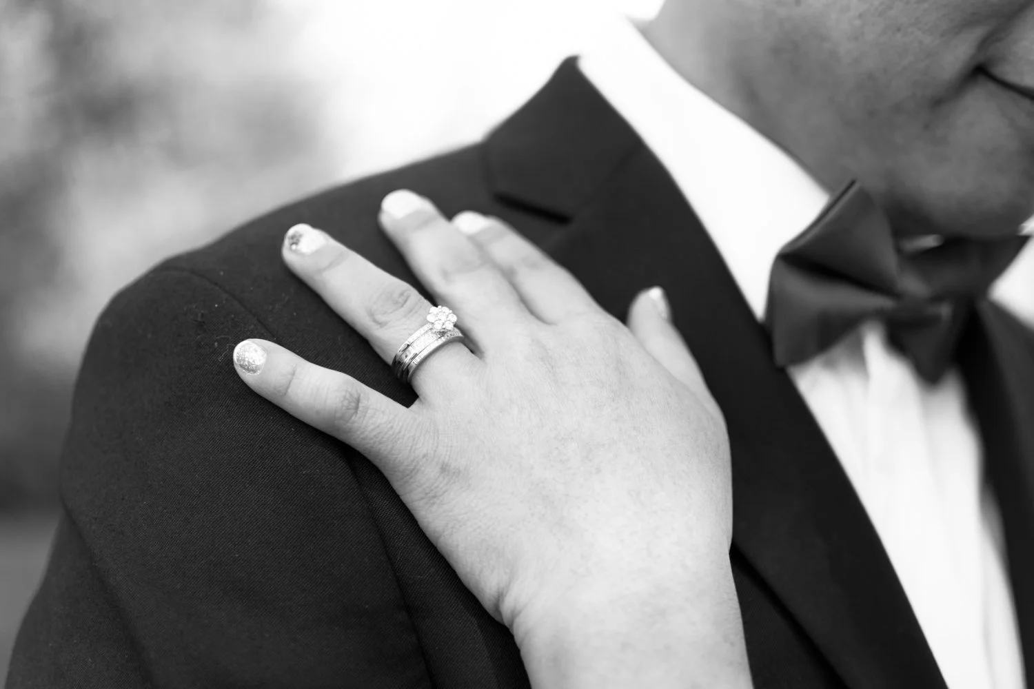 Close-up of a bride's hand with wedding rings on her finger, resting on a groom's shoulder, who is dressed in a tuxedo with a bow tie.