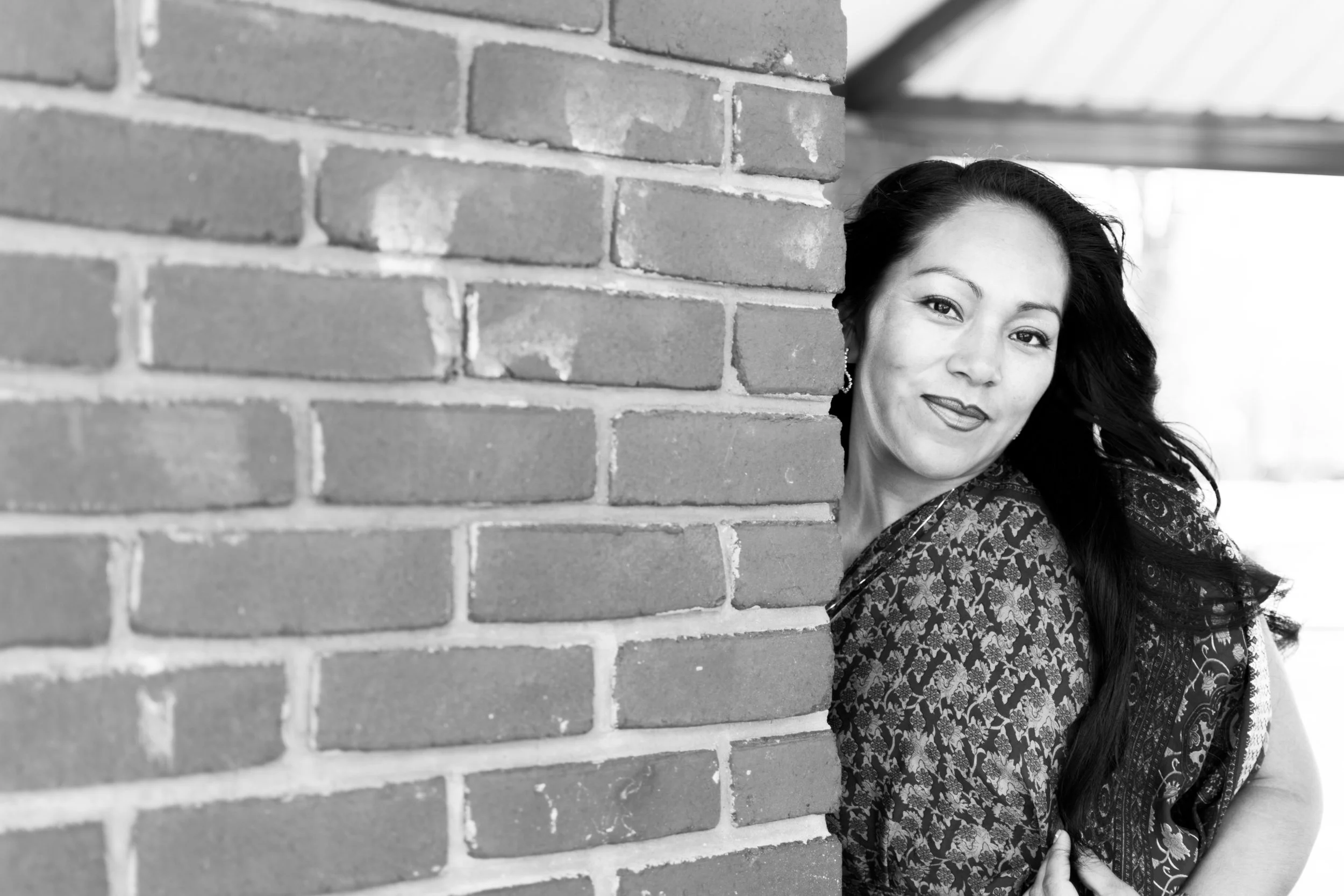 Black and white photo of a woman with long dark hair with a brick wall in the background, smiling and looking at the camera.