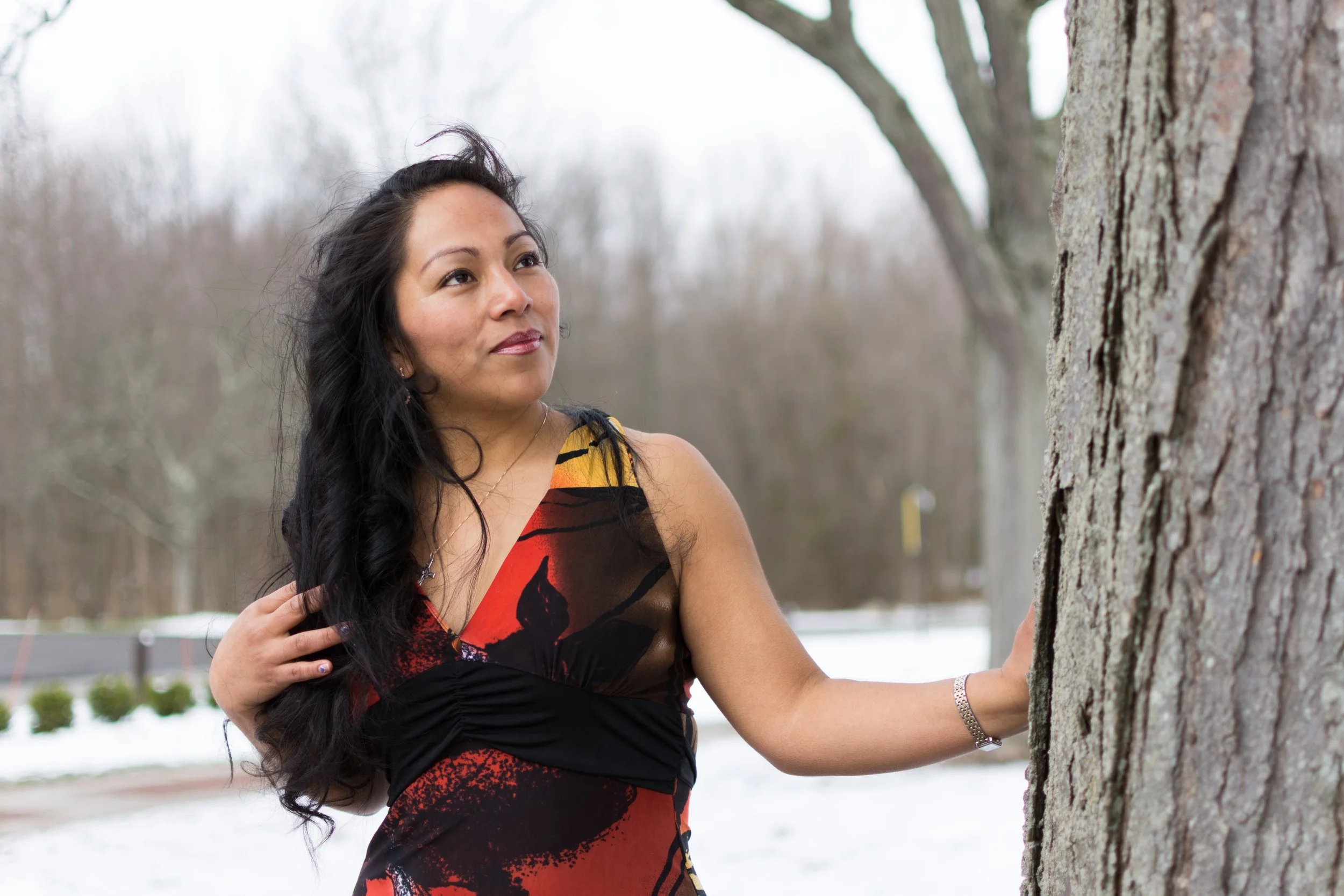 A woman with long black hair, wearing a colorful sleeveless dress, stands outdoors near a tree with a textured bark. She is touching her hair and looking thoughtfully into the distance, with a background of leafless trees and a hint of snow on the gr