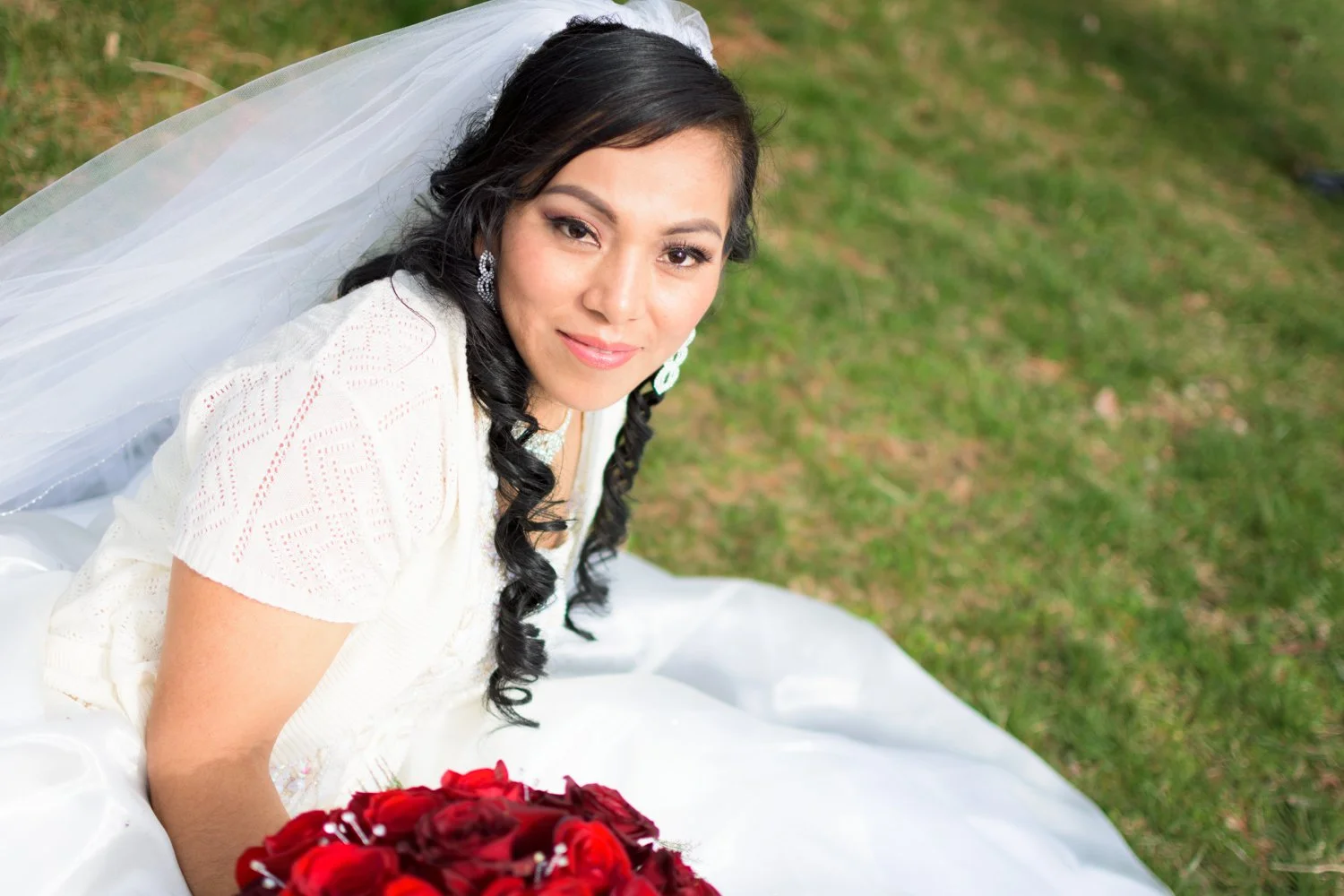 A woman in a white wedding dress sitting on grass, holding a bouquet of red roses, with a veil and earrings, smiling at the camera.