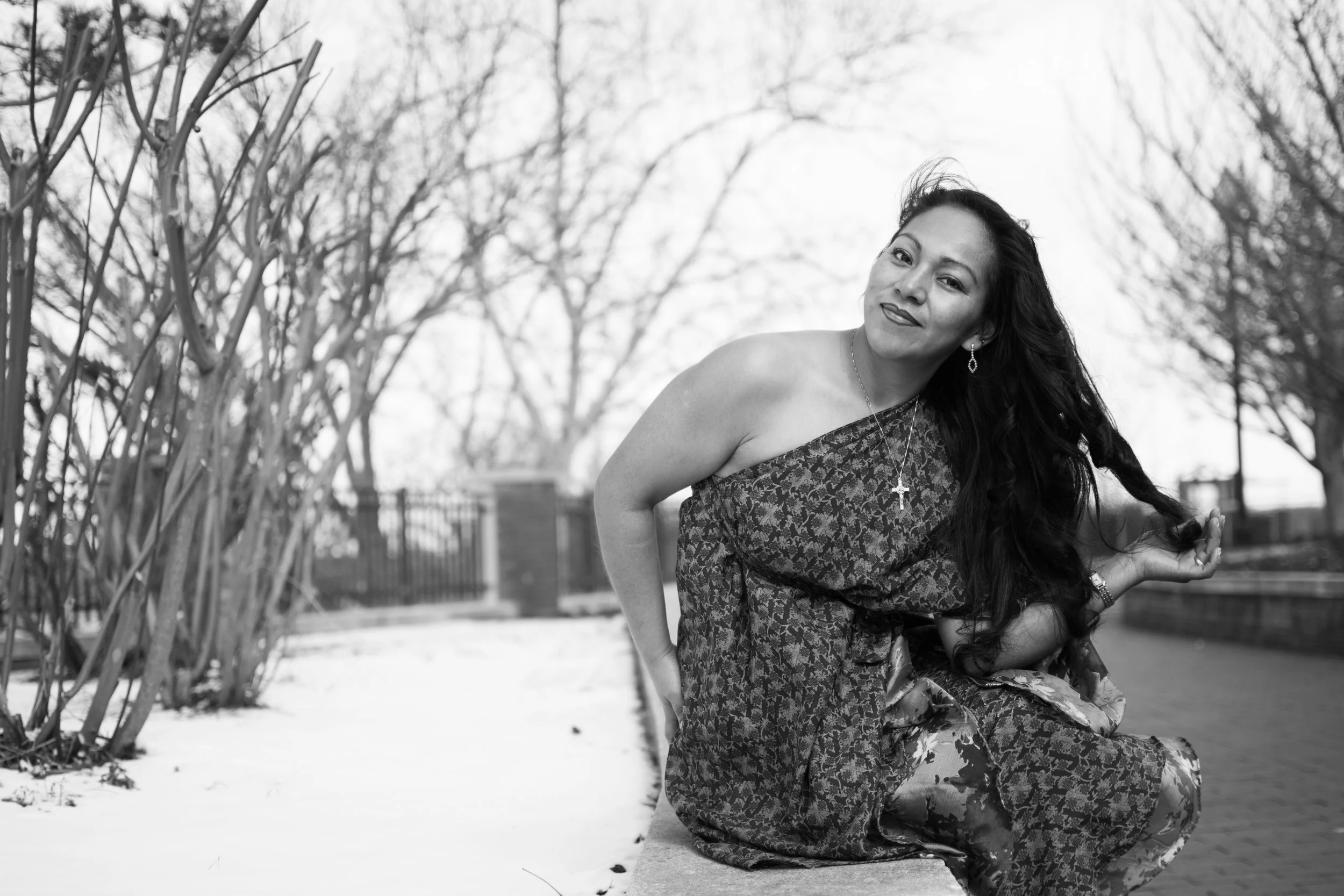 A woman with long dark hair sits outdoors in a park during winter, with bare trees and snow on the ground, wearing a patterned dress and smiling at the camera.