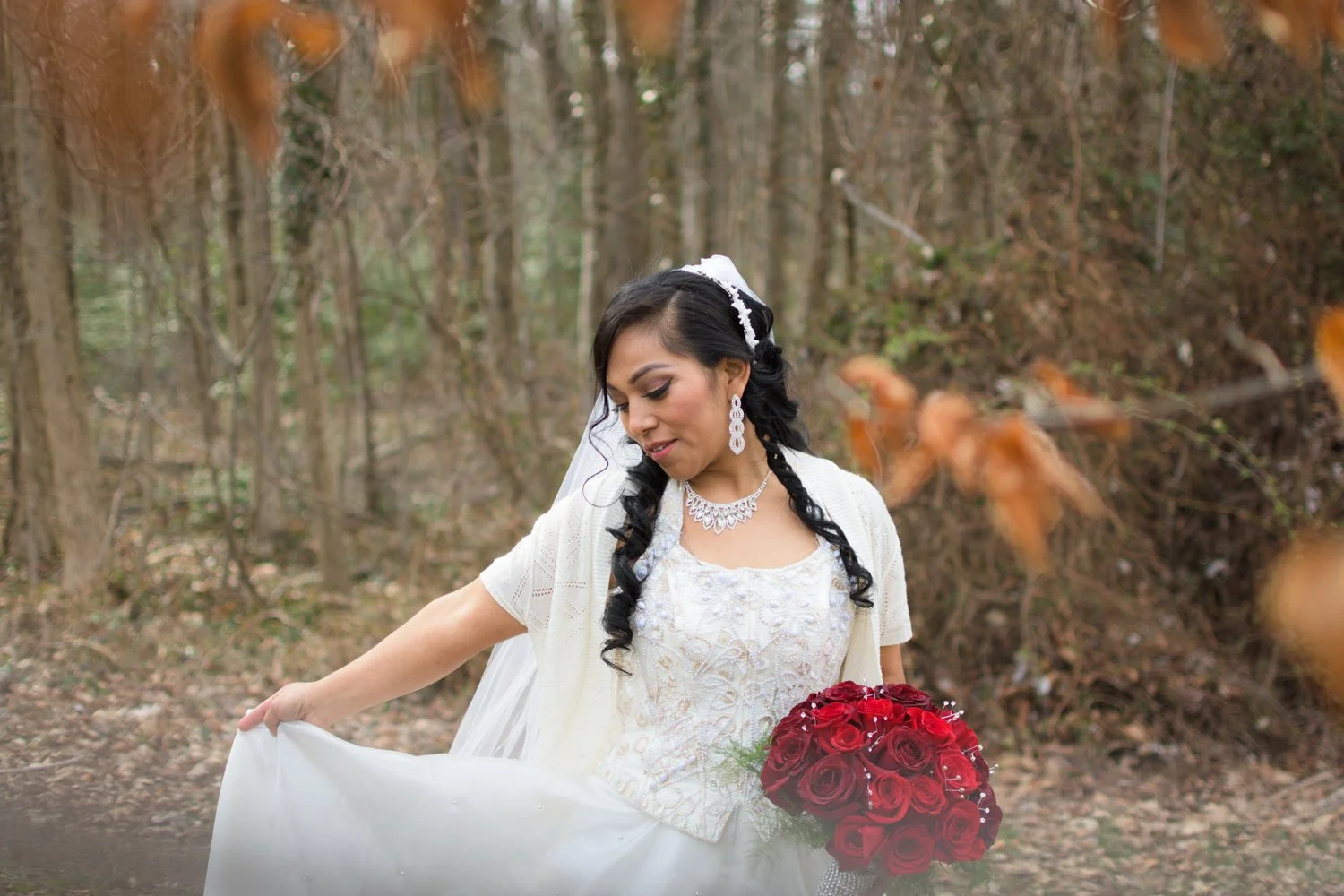 Bride in a white wedding dress holding a bouquet of red roses, standing in a wooded outdoor setting with trees and fallen leaves.