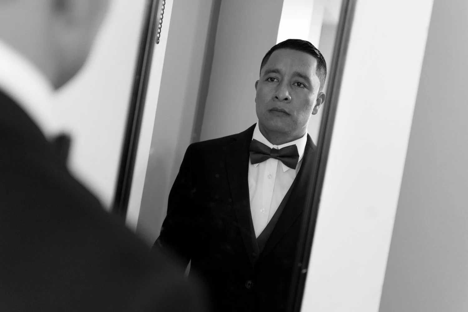 A man in a tuxedo with a bow tie looking at himself in a mirror.