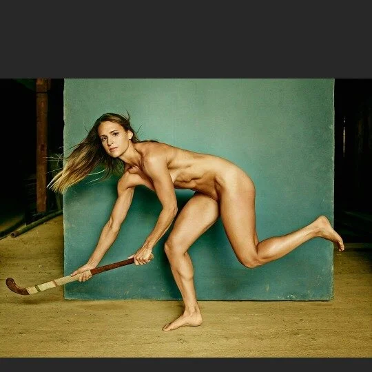 A nude woman with long hair holding a hockey stick, crouched on a wooden floor against a green background.