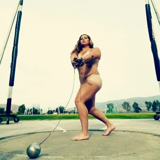 A woman with a tattoo on her hip, holding a shot put, inside a discus throw circle outdoors, with a cloudy sky and trees in the background.