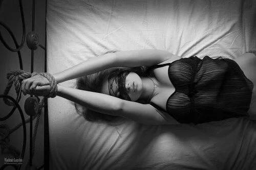 A woman lying in bed with her arms stretched above her head and tied to the bed frame, wearing a black strapless top and a blindfold over her eyes.