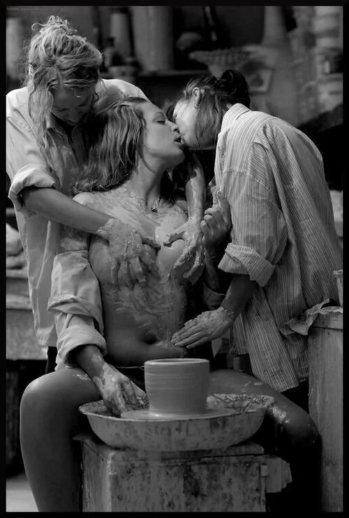 Three women with wet hair and clothes are engaged in an intimate moment, with two women leaning in to kiss and the third woman embracing them from behind, surrounding a central figure who appears to be working on a pottery wheel with a clay vessel.