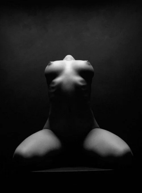 Black and white photo of a reclining nude female figure with her head tilted back, shot from a low angle against a dark background.