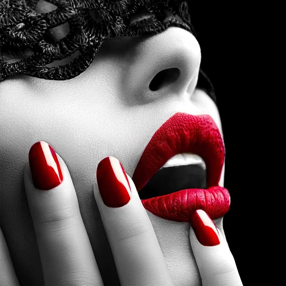 Close-up of a woman's face with red lipstick and red-painted nails, holding her fingers near her lips with a black and white effect on her skin and dark background.