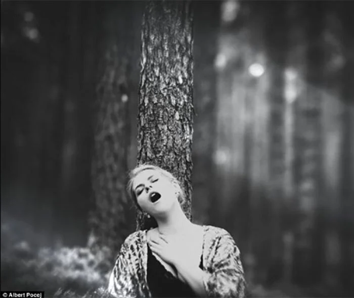 Black and white photo of a woman leaning against a tree in a forest, with her eyes closed and mouth open, holding her neck in a dramatic pose.