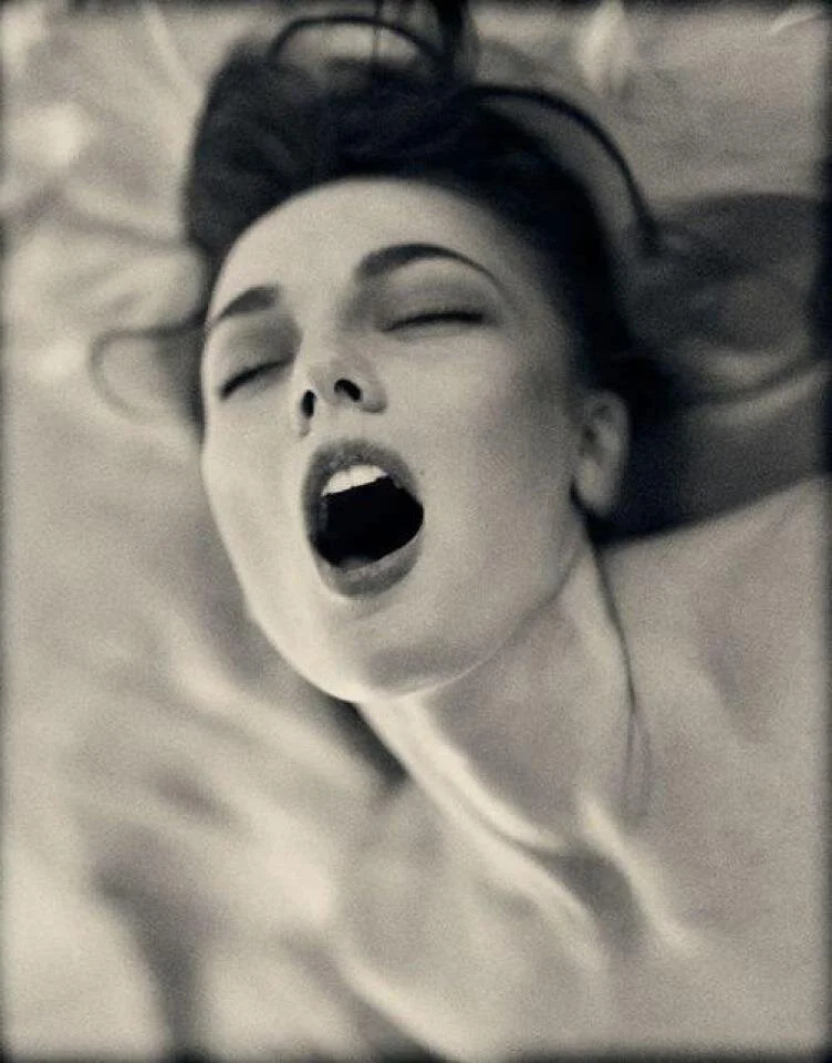 A woman with dark hair lies on her back with her eyes closed and mouth open, possibly yawning or singing.