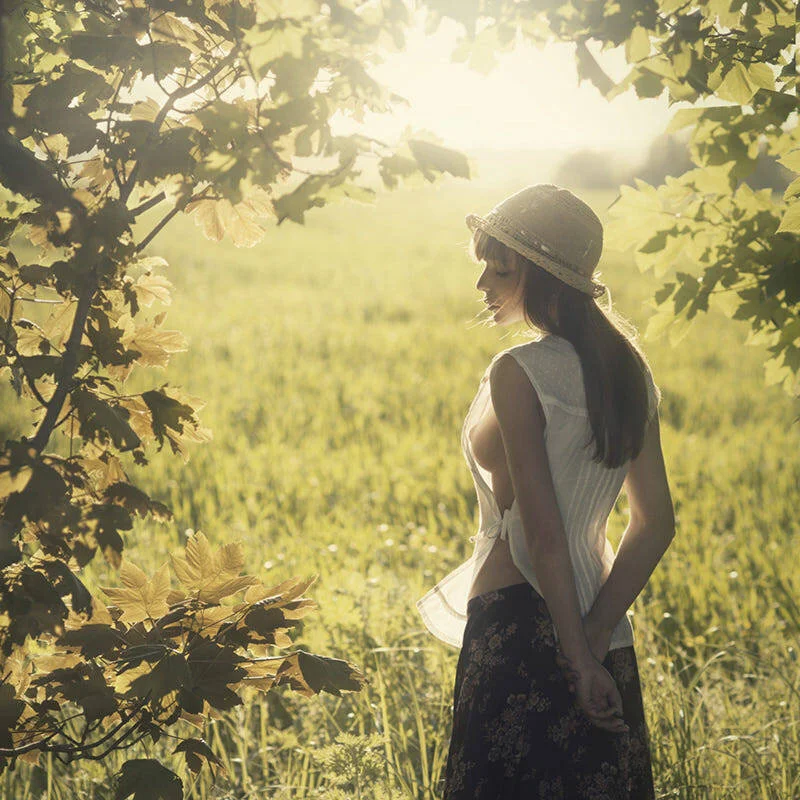 A woman standing in a sunlit field with green grass, wearing a hat, sleeveless top, and patterned skirt, surrounded by trees with large leaves.