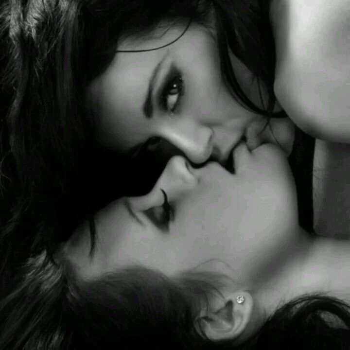 Two women with dark hair lying close together, one kissing the other on the lips in a black and white photo.