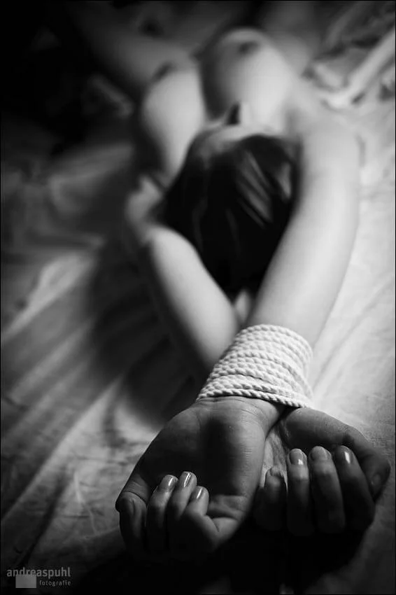 A person lying facedown on a wooden surface with their hands tied behind their back with rope. The focus is on their bound wrists, and their head is turned to the side, partly visible in the background.