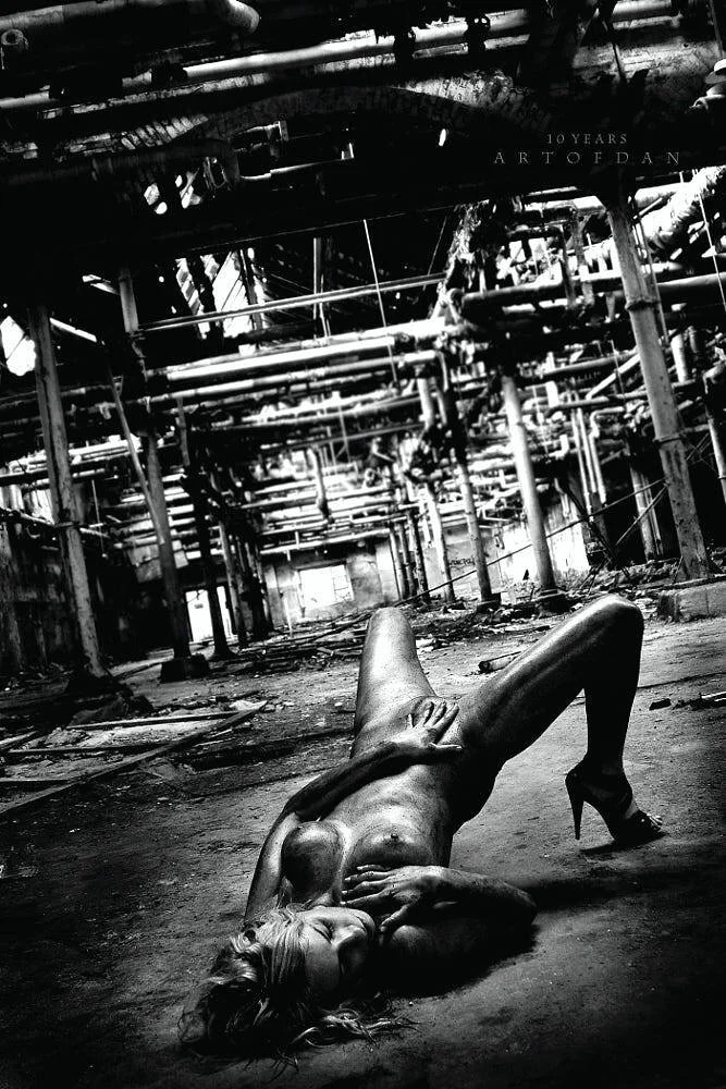 A black-and-white photo of a nude woman lying on the floor of a dilapidated industrial building, with some of her body covered by her arm and her hair spread out, and one leg adorned with a high-heeled shoe. The building is filled with pipes and debr