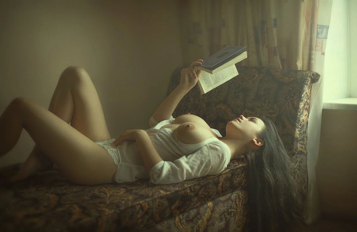 A woman lying on a couch reading a book, with her shirt partially open, in a softly lit room.