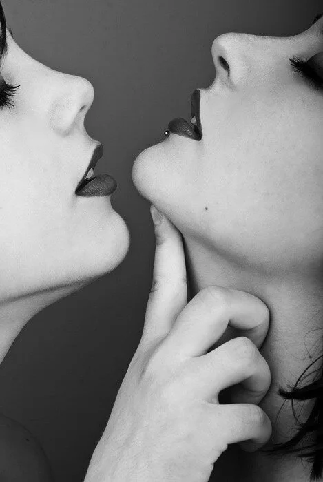 Two women with close-facing faces, one touching the other's chin, both with lips slightly parted and tongues out, in a black and white photograph.