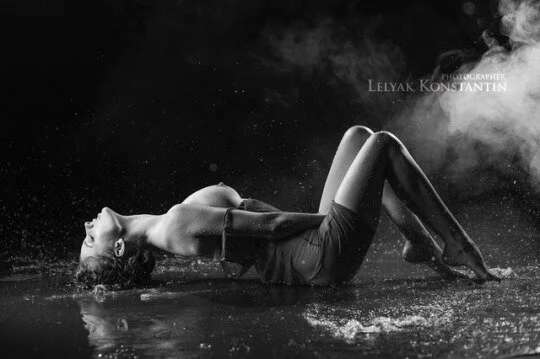 A woman lying on her back in water, arched with her head and shoulders on the surface, in a black and white photograph with smoke.
