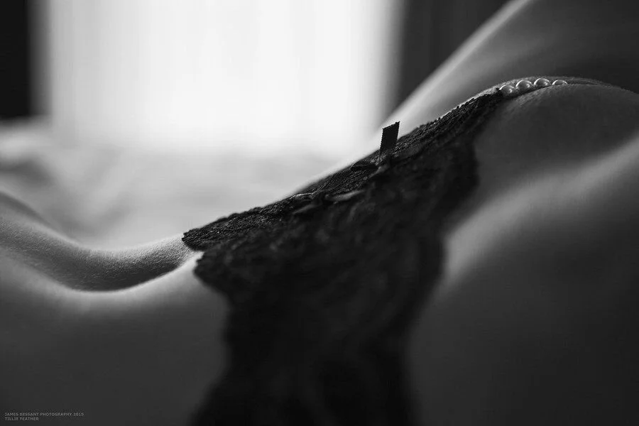 Close-up black and white photograph of a person's midsection, showing a black lace and satin garment with pearl buttons, lying on a bed.