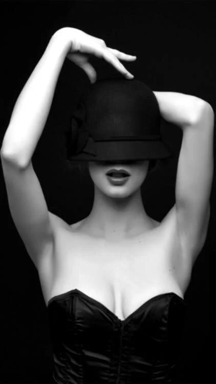 A woman with short hair wearing a black hat that covers her eyes, a strapless black top, and lipstick. She is posing with one arm raised and her hand touching the top hat against a dark background.