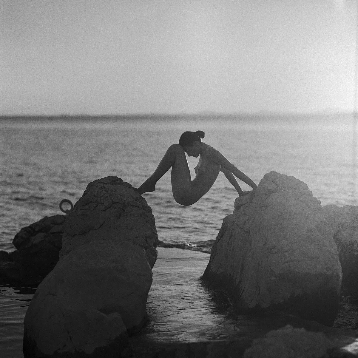 A silhouette of a woman sitting on rocks at the beach, with her legs stretched out and her head bowed, during sunset or sunrise, in black and white.
