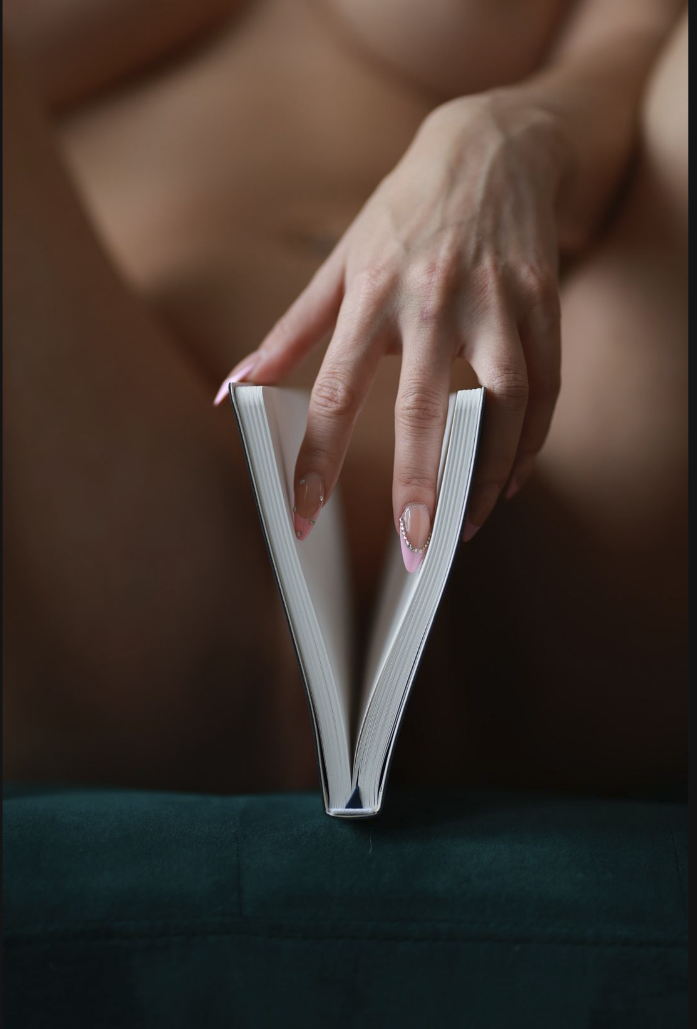 A person's hand with manicured nails holding an open book, with their legs in the background.