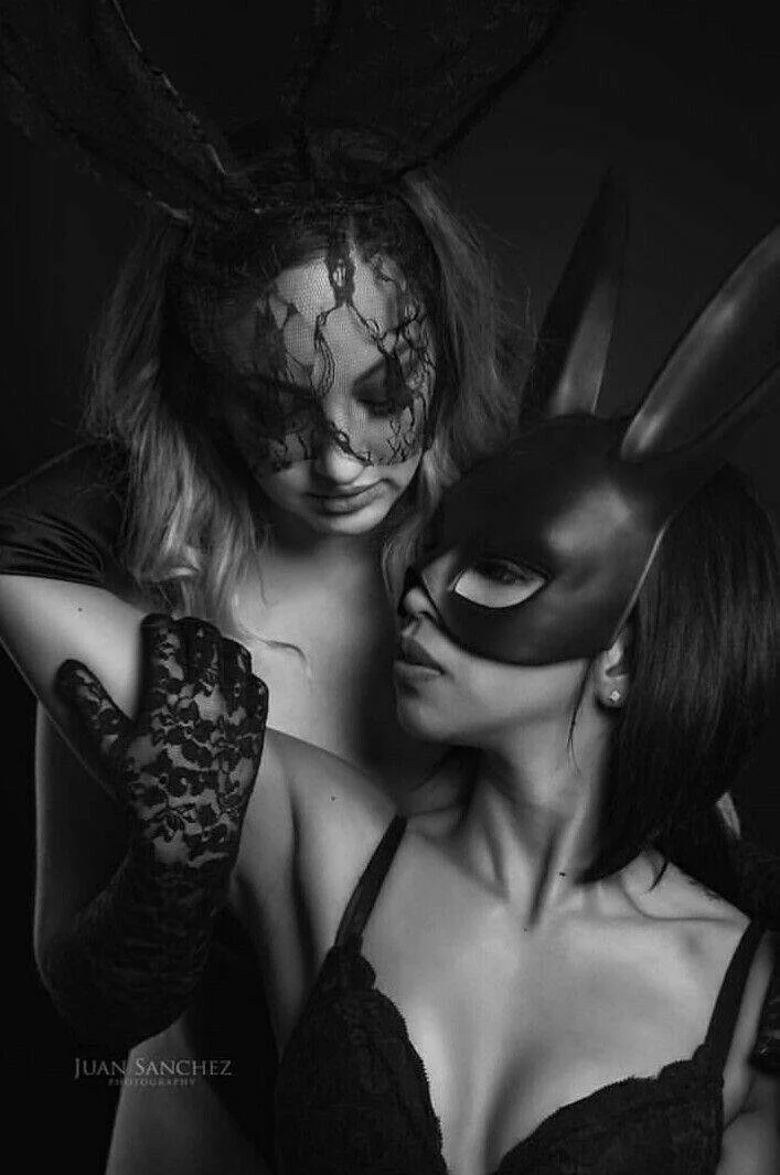Two women dressed in black lingerie and masks, one with rabbit ears, engaging in a close, intimate pose in black and white.