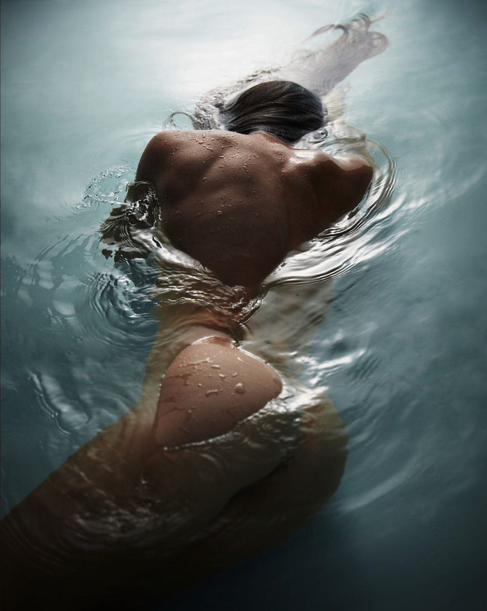 Person swimming underwater, face down, with wet skin and dark hair, in a pool of water.